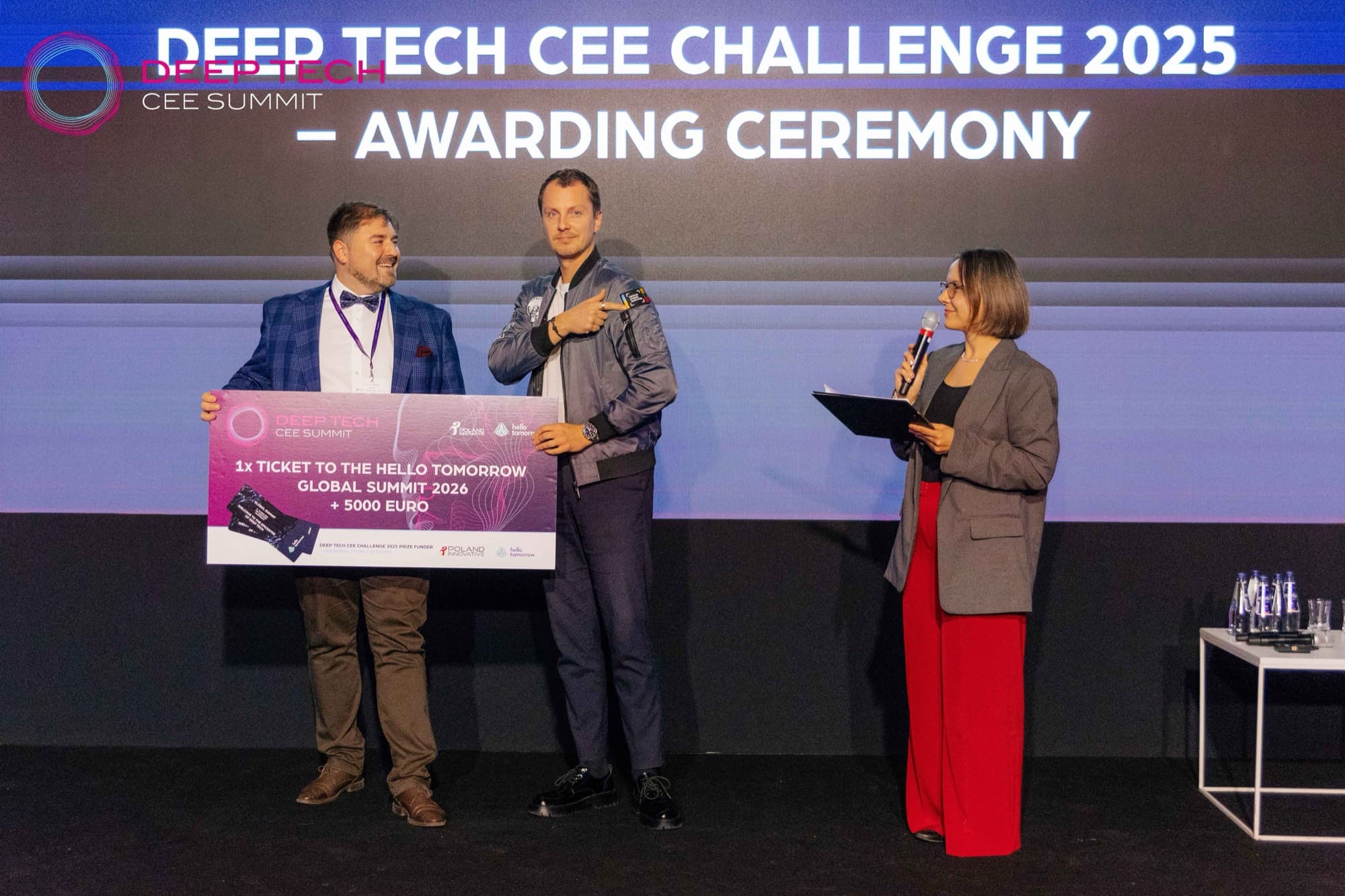 Deep Tech CEE Challenge 2025 awarding ceremony on the Summit stage: a founder holds a cheque for a Hello Tomorrow Global Summit 2026 ticket and €5,000 while the host speaks.